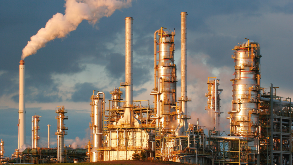 A large, complex oil refinery and chemical processing plant, featuring tall distillation columns and smokestacks, with plumes of steam and smoke rising against a dramatic sunset sky, illustrating industrial infrastructure relevant to Uganda oil refinery investment.