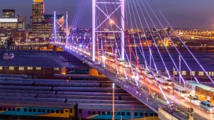 Nighttime cityscape of Johannesburg showcasing the Nelson Mandela Bridge illuminated with traffic flow over active rail lines, highlighting urban infrastructure, mobility, and economic connectivity in South Africa.