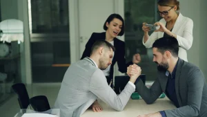 Two businessmen arm-wrestling at a desk while colleagues cheer and take a photo, illustrating an underestimated workforce challenge in corporate competition and team dynamics.
