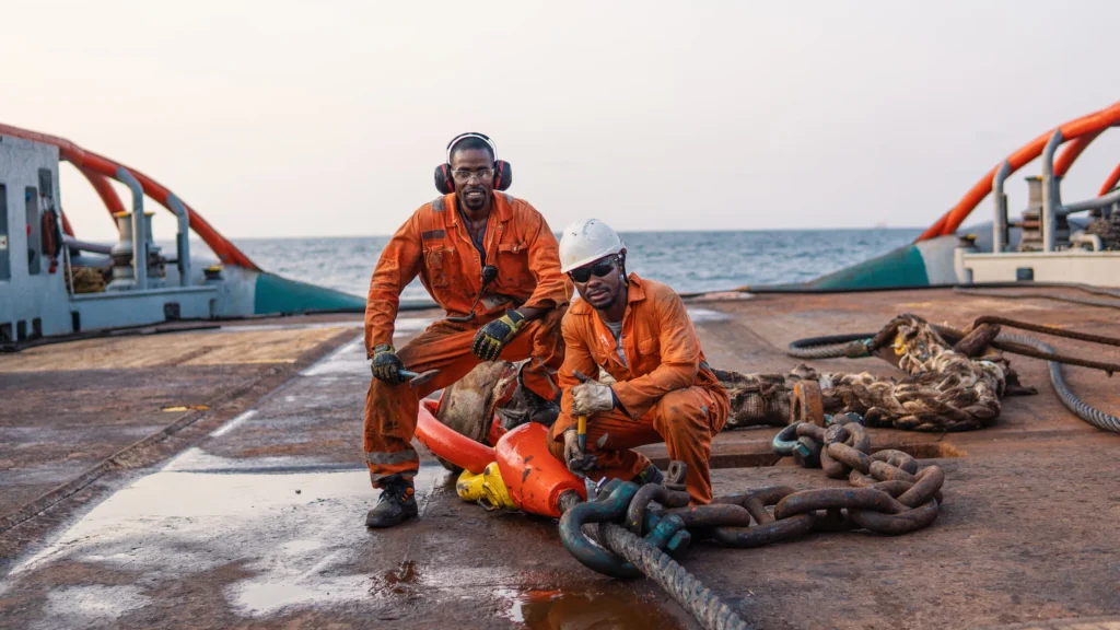 Two offshore maintenance technicians wearing orange coveralls and protective safety gear servicing heavy anchor chains on an offshore platform deck.